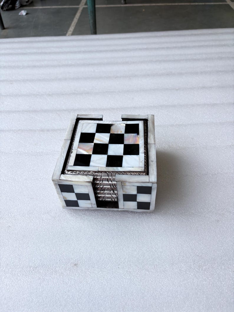 May include: A set of square coasters with a black and white checkerboard pattern on top. The coasters are housed in a matching box with a similar design. The box and coasters have a mother-of-pearl inlay.