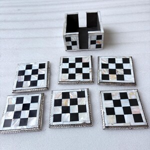 May include: A set of six square coasters with a black and white checkerboard pattern, along with a matching holder. The coasters have a silver-colored border and are made of a mother-of-pearl material.