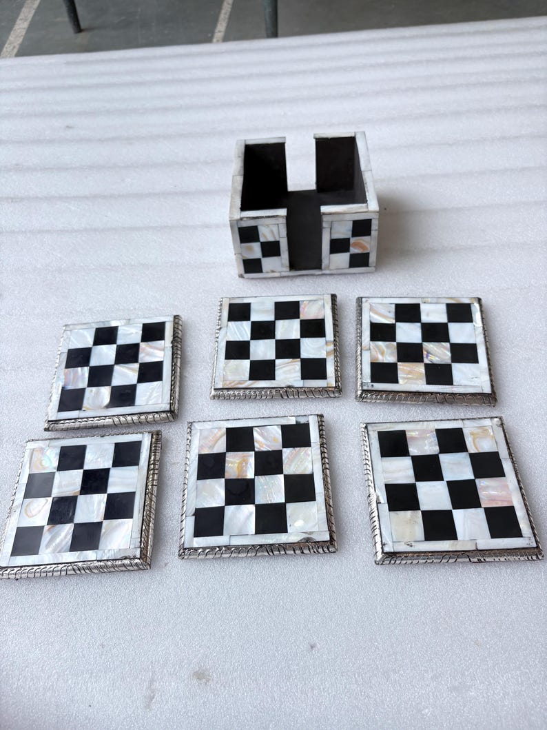 May include: A set of six square coasters with a black and white checkerboard pattern, along with a matching holder. The coasters have a silver-colored border and are made of a mother-of-pearl material.