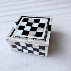May include: A decorative chess-themed box with a checkerboard pattern. The box is made of black and white squares, with a mother-of-pearl effect. The box has a silver trim and is designed to hold chess pieces or other small items.