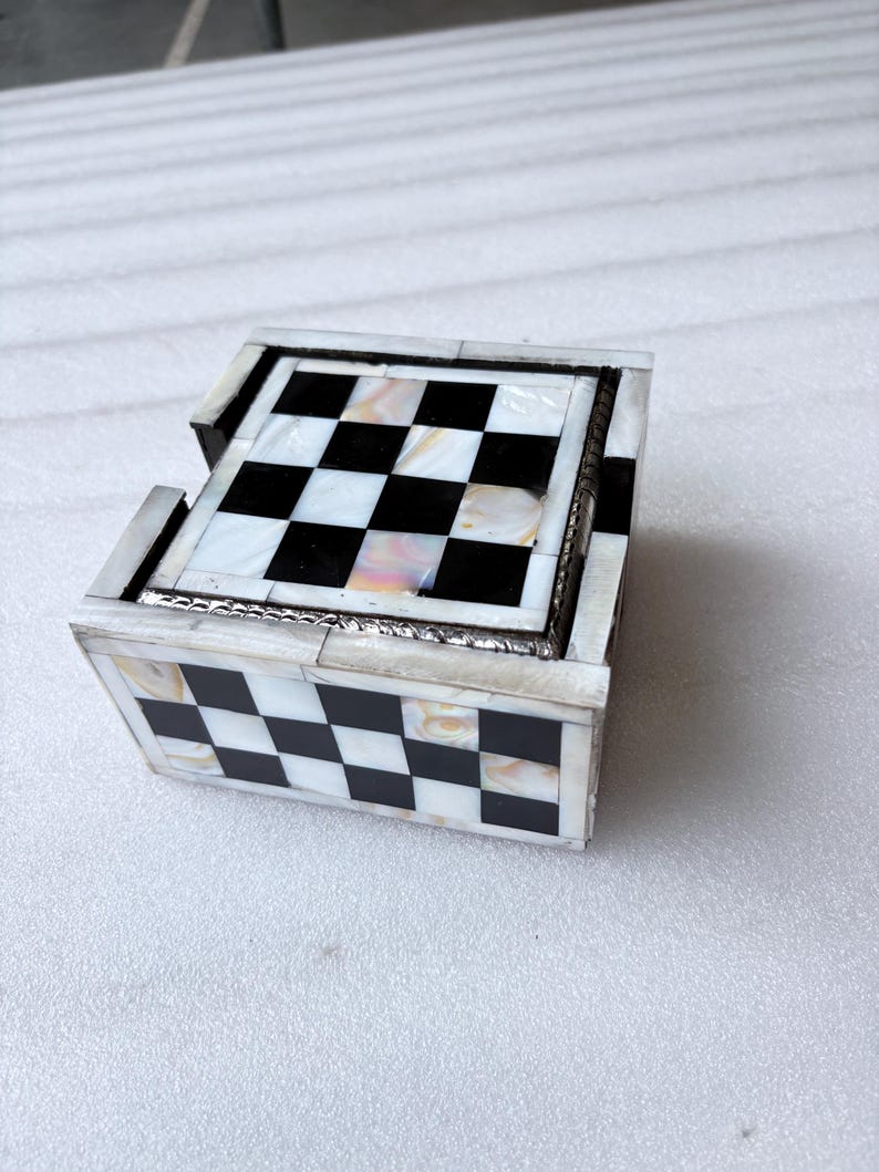 May include: A decorative chess-themed box with a checkerboard pattern. The box is made of black and white squares, with a mother-of-pearl effect. The box has a silver trim and is designed to hold chess pieces or other small items.