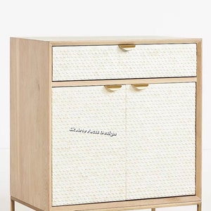 Op de afbeelding: Een witte kast met twee deuren en een lade. De deuren en de lade hebben een getextureerd, wit oppervlak met een subtiel patroon. De kast heeft goudkleurig beslag en poten.