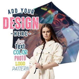 Op de afbeelding: Een vrouw houdt een zwarte paraplu vast met een kleurrijk ontwerp. De paraplu heeft een stadsbeeld en een persoon met een zonnebril erop gedrukt. De tekst "ADD YOUR DESIGN HERE" staat in roze en blauwe letters. De tekst "Any Text COLOR PHOTO LOGO PATTERN" staat in kleurrijke letters.