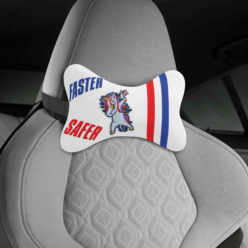 Puede incluir: Coj&iacute;n blanco para reposacabezas de coche con las palabras "FASTER" y "SAFER" en azul y rojo. El coj&iacute;n presenta un unicornio de dibujos animados con una melena y cola rojas, blancas y azules. Hay rayas rojas y azules en el lado derecho.