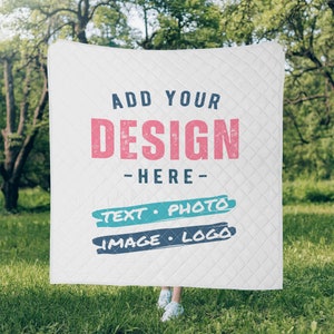 Op de afbeelding: Witte gewatteerde deken met de tekst "ADD YOUR DESIGN - HERE". Onder de tekst staan de woorden "TEXT • PHOTO" en "IMAGE • LOGO" in een turquoise en marineblauw ontwerp. De deken wordt vastgehouden voor een groene, grasrijke achtergrond.
