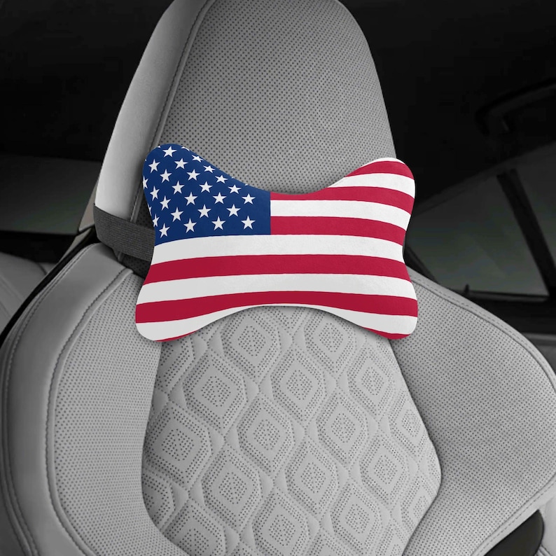 Puede incluir: Un coj&iacute;n para reposacabezas de coche con el dise&ntilde;o de la bandera estadounidense. El coj&iacute;n tiene un campo azul con estrellas blancas y rayas rojas y blancas. Se coloca en un asiento de coche gris.
