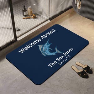 May include: A rectangular navy blue doormat with the text "Welcome Aboard" and "The Sea Jones Surry, VA" in white. A light blue marlin fish graphic is in the center. Black sandals are on the floor.