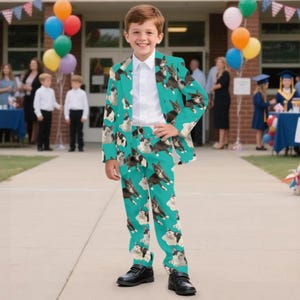 May include: A boy wearing a teal suit patterned with Boston Terrier dogs. The suit includes a jacket and matching pants. He is also wearing a white dress shirt and black shoes. Balloons and people are in the background.