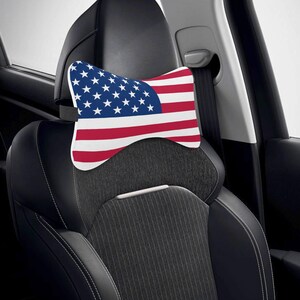 Puede incluir: Un coj&iacute;n para reposacabezas de coche con el dise&ntilde;o de la bandera estadounidense. El coj&iacute;n tiene un fondo blanco con rayas rojas y blancas y un campo azul de estrellas. Se coloca en un asiento de coche negro.