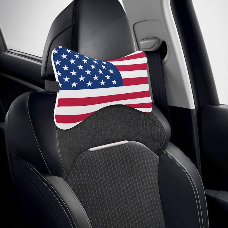 Puede incluir: Un coj&iacute;n para reposacabezas de coche con el dise&ntilde;o de la bandera estadounidense. El coj&iacute;n tiene un fondo blanco con rayas rojas y blancas y un campo azul de estrellas. Se coloca en un asiento de coche negro.