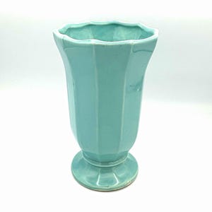 May include: A light turquoise ceramic vase with a glossy finish, featuring a fluted, vertical panel design and a scalloped rim. The vase stands on a circular pedestal base, showcasing a smooth, uniform color throughout its entire structure.