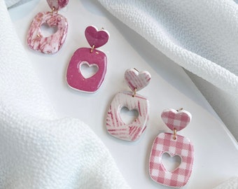 May include: A set of four pink and white heart-shaped earrings. Each earring features a different pattern, including gingham, marble, and floral designs. The earrings are made of polymer clay and have gold-tone hooks.
