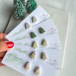 Peut inclure: Un ensemble de six paires de boucles d'oreilles en forme de sapin de Noël. Les boucles d'oreilles sont en pâte polymère et sont disponibles en trois couleurs : vert, blanc et beige. Les boucles d'oreilles sont présentées sur des cartes avec le nom de l'entreprise "BGLAMBOUTIK" et des informations sur le site Web.