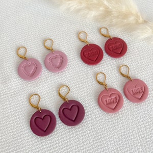 Peut inclure: Six paires de boucles d'oreilles en forme de cœur avec des crochets dorés. Les boucles d'oreilles sont en pâte polymère et se déclinent en trois couleurs : bordeaux, rose et rose clair. Les cœurs ont des motifs différents, notamment un cœur simple, un cœur avec une bordure et des cœurs avec les mots « XOXO » et « LOVE » en relief.
