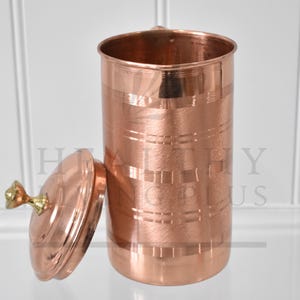 May include: A copper-coloured cylindrical container with a lid. The container has horizontal and vertical etched lines. The lid is off to the side, revealing the interior. The lid has a gold-coloured knob. The text "HEALTHY PLUS" is visible on the side of the container.