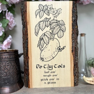 May include: A wooden sign with a black and white illustration of a potato plant. The sign reads "Po Täy-Toës" and includes the text "boil em', mash em', stick em' in a stocca".
