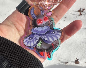 May include: A clear acrylic keychain featuring a cartoon moth with large red eyes, purple wings, and a pink heart. The moth is wearing a fluffy purple collar and red shoes. The keychain has a rose gold metal ring.