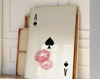 May include: A large framed print of the Ace of Spades playing card with a pair of pink lipstick kiss marks. The artwork leans against a white wall in a room with hardwood floors.