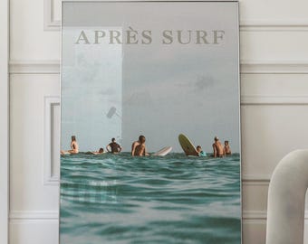 May include: A framed print featuring a serene ocean scene with surfers and surfboards. The text "APRÈS SURF" is displayed at the top. The image is in a silver frame and is displayed against a white wall and a wooden floor.