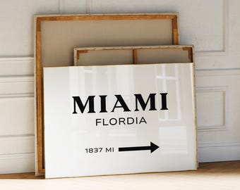 May include: A framed white sign with the words "MIAMI FLORDIA" in black, with an arrow and the text "1837 MI" pointing right. The sign is propped against a wall with two other framed canvases.