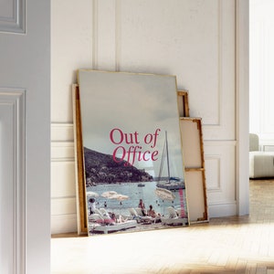 May include: A framed poster with a photograph of a beach scene with a sailboat and people on the beach. The poster has the text "Out of Office" in pink letters.