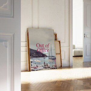 May include: A framed print with a photo of a beach scene with a sailboat and people on the beach. The text "Out of Office" is printed in pink on the print.