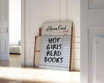 May include: A framed library card art print with the text "HOT GIRLS READ BOOKS" in bold black letters. The print is propped against a wall in a room with a wooden floor and white walls.