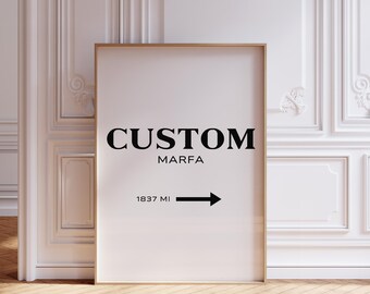 May include: A framed art print with the word "CUSTOM" above "MARFA" in black font. Below, an arrow points right with "1837 MI" in black. The print has a gold frame and is set against a white wall.