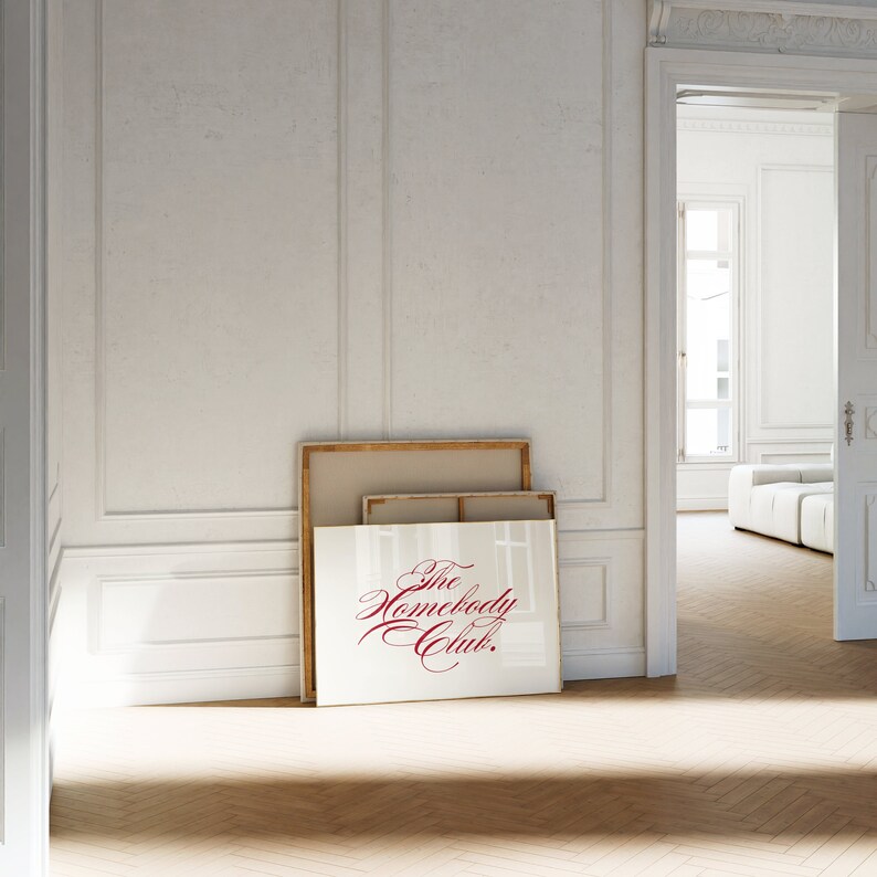 May include: Two framed pictures leaning against a white wall. The top picture is a plain canvas, and the bottom picture is a gold frame with the words "The Homebody Club." in red cursive.