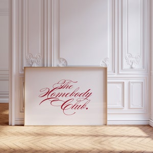 May include: A gold framed print with a white background and the text "The Homebody Club." in red cursive lettering.