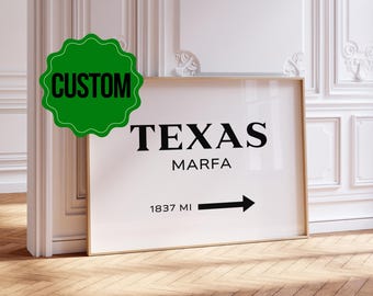 May include: A framed print with the word "TEXAS" above "MARFA" in black font. Below, an arrow points right with "1837 MI". A green "CUSTOM" sticker is in the upper left corner. The print has a gold frame and is displayed on a wooden floor.