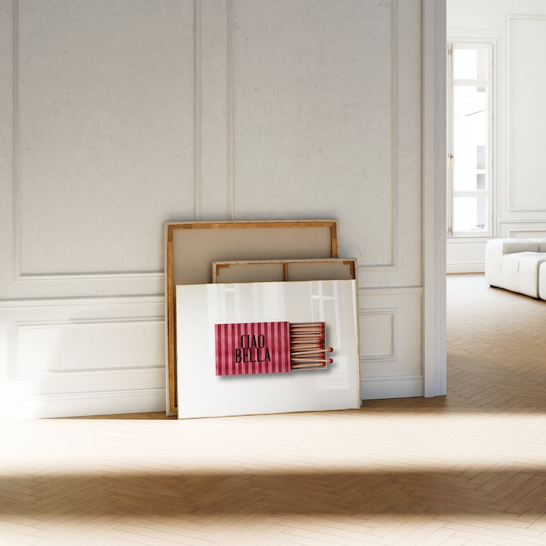 May include: A white framed print with a red and white striped matchbox with the text "Ciao Bella" on it. The print is leaning against two other canvases.
