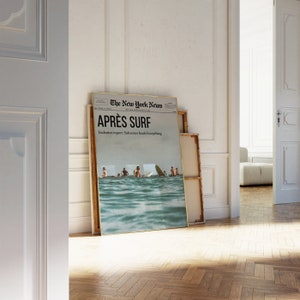 May include: A framed poster leaning against a wall, featuring a newspaper-style design with the headline "APR&Egrave;S SURF." The image shows surfers in the ocean. The text "Exclusive report: Saltwater heals Everything" is also visible. The frame has a gold border.