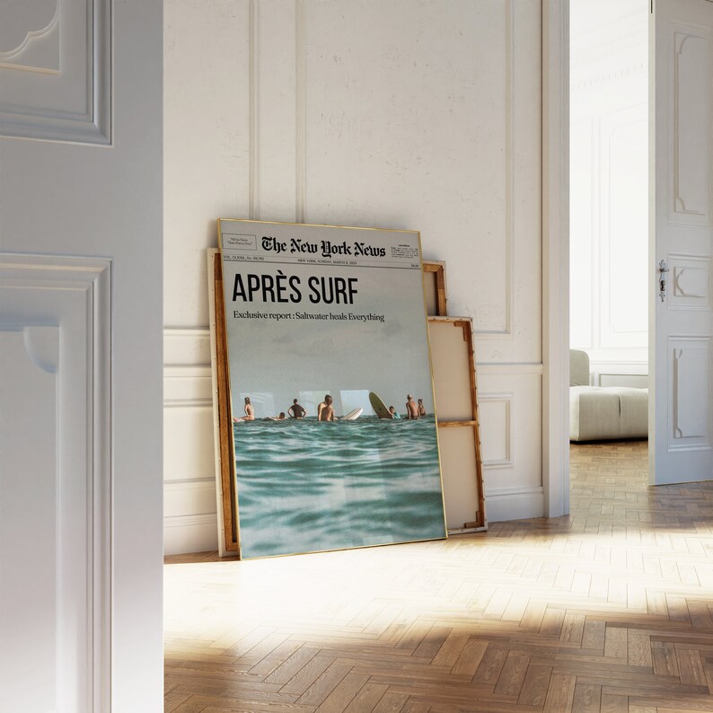 May include: A framed poster leaning against a wall, featuring a newspaper-style design with the headline "APR&Egrave;S SURF." The image shows surfers in the ocean. The text "Exclusive report: Saltwater heals Everything" is also visible. The frame has a gold border.
