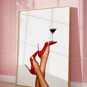 Op de afbeelding: Een ingelijste print van een paar rode hakken die een martini-glas met een donkere vloeistof vasthouden. De achtergrond is wit.