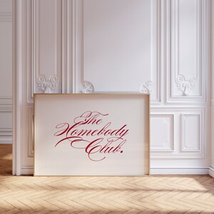 May include: A gold framed print with a white background and the text "The Homebody Club." in red cursive script.