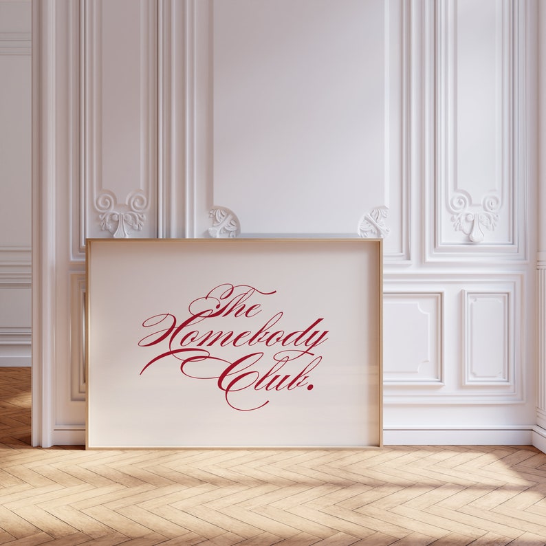 May include: A gold framed print with a white background and the text "The Homebody Club." in red cursive script.