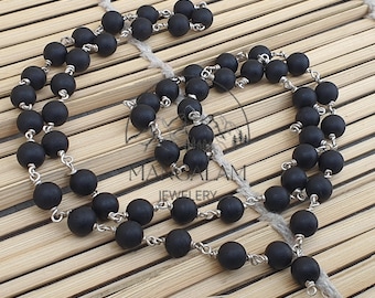 May include: A black beaded necklace with silver-toned metal links, displayed on a light brown, textured surface. The necklace is composed of numerous round, matte black beads. The design includes a central pendant and a matching set of beads.