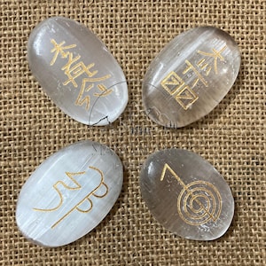 May include: Four oval selenite stones with gold symbols and characters. The stones range in colour from white to translucent grey. The symbols are etched into the surface. The background is a tan woven fabric.
