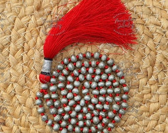 May include: A handcrafted necklace featuring a circular arrangement of silver-colored beads interspersed with red beads. A vibrant red tassel with a silver and black accent adds a striking contrast. The necklace rests on a textured, woven background.