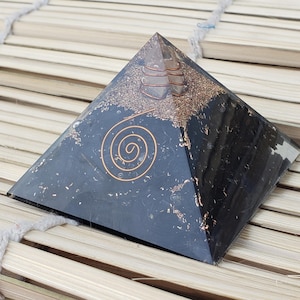 May include: A black orgone pyramid with a copper spiral design on the front. The pyramid has a clear top with copper and other materials inside. The pyramid is sitting on a bamboo surface.