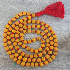 Pode incluir: Um colar mala com contas amarelas enfiadas num cordão vermelho, com uma borla vermelha brilhante. As contas são de uma cor amarela quente, e o cordão e a borla vermelhos criam um contraste marcante. Este colar é provavelmente usado para meditação ou práticas espirituais.