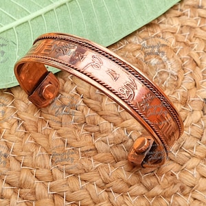May include: Copper cuff bracelet with etched patterns and text. The bracelet has a hinged design with a clasp closure. The surface has a textured appearance, and the bracelet is displayed on a woven background.