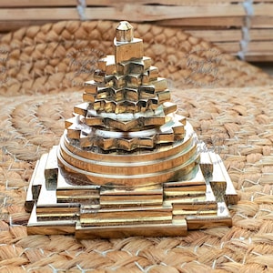 May include: A gold-colored, tiered geometric sculpture with a pyramid-like shape. The sculpture has multiple layers with star-shaped and rectangular elements. The piece is displayed on a woven, textured surface.