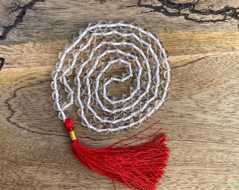 Op de afbeelding: Een heldere kwarts kristallen mala ketting met een felrode kwast. De kralen zijn in een cirkelvormig patroon gerangschikt, waardoor een spiritueel en decoratief item ontstaat. De kwast is gemaakt van rode draden.