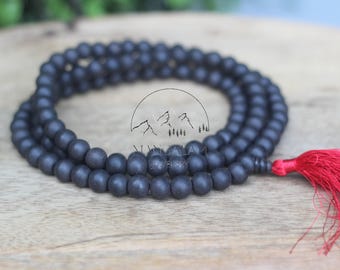 May include: A dark grey beaded mala necklace with a red tassel. The beads are round and strung together, forming a looped design. The necklace is displayed on a wooden surface.