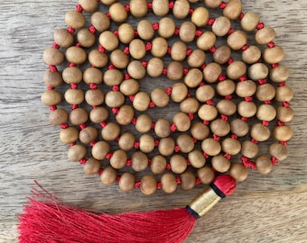 May include: A wooden mala bead necklace with 108 light brown beads strung on red thread. The necklace has a red tassel with a gold and black accent at the end. The beads are smooth and round, and the tassel is full and flowing.