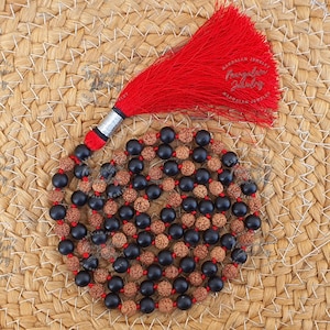 May include: A necklace featuring black and brown beads, and a red tassel. The beads are strung with red thread. The tassel has a silver and black accent. The necklace rests on a woven, tan surface.