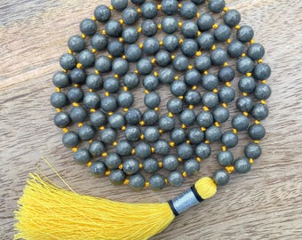 May include: A gray pyrite gemstone mala necklace with a bright yellow tassel. The beads are strung on a gold-colored cord. The tassel is attached to the end of the necklace with a silver and black accent. The necklace is displayed on a wooden surface.