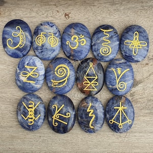 May include: Twelve oval sodalite stones, each with a deep blue hue and white veining. Gold symbols adorn each stone. The stones are displayed on a light brown wooden surface.